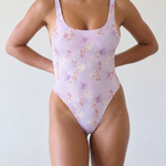 A woman modeling a light lavender one-piece swimsuit with a delicate pastel floral pattern, standing against a plain off-white background. Kaniela Print - Julia One Piece - Inherited Collection